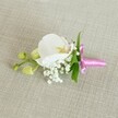 A delicate orchid boutonniere or corsage is laid horizontally on a softly textured, light beige linen-style fabric, the neutral background allowing every floral detail to stand out with quiet elegance. At the heart of the design sits a single, pristine white Phalaenopsis orchid, its broad, velvety petals perfectly smooth and gently curved, framing a centre touched with soft yellow and the faintest whisper of pale pink. Nestled closely around the base of the orchid are tiny, cloud-like clusters of white baby's breath, each miniature bloom creating a misty, ethereal halo that softens the overall look. Slender, vibrant green leaves peek out from behind the flowers, adding a fresh, natural contrast that suggests a walk through one of Grove Park's leafy side streets on a calm morning. To one side, several unopened lime-green orchid buds rest along a thin stem, their rounded shapes hinting at blossoms yet to unfurl and adding a subtle sense of movement and growth. The stems of the flowers and foliage are neatly bound together with a slim, bright fuchsia pink satin ribbon, tied into a small, precise bow whose glossy sheen catches the gentle, even light. The pop of vivid pink against the creamy orchid and white gypsophila gives the piece a modern yet timeless feel, ideal for a groom's lapel, a prom date, or a special guest at a Grove Park celebration. The overall impression is one of refined simplicity and quiet joy, like the moments just before guests arrive for a wedding or formal gathering nearby, when everything is prepared and anticipation hangs sweetly in the air.