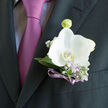 The photograph reveals a single white orchid boutonniere pinned neatly to the lapel of a dark suit, a study in balance, texture, and quiet celebration. The orchid is immaculate, with broad, smooth petals that radiate outwards in a gentle curve, giving the bloom a sculpted, almost porcelain quality. At the very centre, subtle hints of warm ivory and pale blush add depth without disturbing the overall purity of the flower. Encircling it, slender silvery-green leaves and delicate lilac blooms create a mist-like halo, softening the transition between the crisp suit fabric and the organic forms of the flowers. Tiny white buds and gypsophila peep through, like scattered stars in a twilight sky. The stems are wrapped in a narrow violet ribbon, tightly secured and finished with enviable neatness, its sheen catching just enough light to echo the purple-lilac tie worn underneath. The dark jacket provides a rich backdrop, the weave of the fabric visible enough to suggest fine tailoring and a special occasion-perhaps a Grove Park ceremony or reception near Chiswick House, with guests arriving from nearby Strand-on-the-Green. The overall impression is of something prepared with care in a local studio, then carefully carried and pinned in place just before photographs begin. You can imagine the soft murmur of congratulations, the rustle of suits and dresses, and the almost imperceptible floral scent-fresh, clean, and lightly sweet. This boutonniere is not merely decorative; it's a thoughtful finishing touch that reflects the wearer's style and the emotional weight of the day, captured in one luminous orchid and a few perfectly chosen accents.
