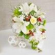 Displayed on a flawless white table, a sophisticated floral arrangement glows gently against a soft-beige textured background, like a carefully styled wedding centerpiece ready for a reception in Grove Park. The flowers are arranged in a round white wicker basket, its finely woven texture introducing a natural, handcrafted note that complements the elegant blooms. Dominating the design are several large white lilies, fully open and trumpet-shaped, their creamy petals curling outward with subtle speckles near the center and stamens dusted in pale yellow pollen, hinting at a rich, sweet fragrance. Close to the upper center of the bouquet, a single cream to pale yellow rose unfurls in a tight swirl, offering a warm, romantic focal point. From the right side, white phalaenopsis orchids spill down like a delicate waterfall, each bloom with wide, softly rounded petals that lend the arrangement a teardrop or cascade shape often chosen for bridal tables along the Thames path. Bright green pom-pom chrysanthemums nestle between the lilies and orchids, their rounded, textured forms adding playful contrast. Threads of feathery greenery and broader, glossy leaves weave through the bouquet, giving it structure and a lush garden feel. Tiny white baby's breath blossoms scatter lightness throughout, while clusters of glossy red berries punctuate the white and green palette with vibrant flashes of colour. Resting to the left on the tabletop are two white wicker spheres of different sizes, their open, interlaced design echoing the basket and providing a modern decorative touch. The overall impression is one of polished elegance and fresh, fragrant beauty, the kind of refined table arrangement Grove Park Florist might create for weddings, anniversaries, and intimate celebrations across the local area.