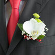 Pinned neatly to a dark suit jacket, this boutonnière showcases the kind of thoughtful floral design that would feel perfectly at home at a Grove Park wedding reception. At the focal point is a pristine white orchid, its petals smooth, velvety, and gently overlapping in a natural, open shape. A hint of warm yellow at its centre adds depth and a touch of softness, drawing attention to the flower's elegant form. Nestled behind, there are lime-green button chrysanthemums, compact and spherical, their dense petals forming textured domes of colour that evoke the fresh, grassy banks along the Thames near Chiswick. Interspersed around the main blooms, delicate stems of baby's breath extend outward, each carrying clusters of minute white flowers that appear almost like fine lace or a light mist. These tiny details lend a sense of weightlessness and romance to the design. Framing the flowers, glossy dark green leaves offer structure and contrast, their clean lines standing out crisply against the darker weave of the lapel. At the base, a slim ribbon in deep red is carefully wrapped and tied, harmonising with the matching red tie worn beneath and adding a subtle note of passion to the otherwise calm palette. The overall composition feels balanced and graceful, like a quiet promise held close to the heart. This boutonnière would suit a groom, best man, or honoured guest, accompanying them as they move from the ceremony to photographs in nearby Grove Park, and finally to an evening of toasts and dancing under soft, golden lighting.