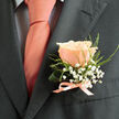 Pinned with precision to a deep grey suit lapel, this peach-pink rose boutonnière embodies understated romance and expert floral craftsmanship. The single bloom is the focal point: petals layered tightly toward the heart and loosening at the edges, their surface catching soft daylight to reveal creamy peach tones that graduate into blush pink. Around the rose, scattered sprays of tiny white gypsophila create a lacy halo, while slender strands of pale green foliage introduce a subtle asymmetry that feels deliberately organic rather than rigid. The small satin ribbon at the stem is a tender salute to the tie's warm salmon hue, its lustrous loop neatly bound so the entire piece sits flat and secure against a light dress shirt patterned with a faint dark grid. The charcoal jacket provides a structured, elegant frame-its smooth weave setting off both the softness and the meticulous detail of the floral work. I prepared pieces like this for ceremonies near Grove Park station and the Nature Reserve, where couples often ask for garden-inspired touches that nod to quiet neighbourhood charm; the boutonnière's scent is a whisper of fresh-cut rose, the texture velvety and intimate. This accessory reads as both a refinement of menswear and a small poem of tenderness-perfect for a groom, best man, or guest seeking a subtle but meaningful floral accent for a wedding or formal evening in and around Grove Park.