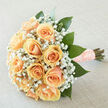 This arrangement reads like a soft promise: apricot-peach roses clustered into a compact, rounded bouquet with petals that curl inward in velvety layers, each bloom carefully selected for size and tone. Surrounding them is an ethereal cloud of white gypsophila - tiny, light-filled blossoms that weave through the roses and create a delicate counterpoint. Rich emerald foliage frames the composition, the deep green leaves shaped to cradle the flowers and accentuate the pastel warmth. The stems are neatly bound in a textured peach ribbon, the wrap secured with pearl-headed pins that provide a tactile, old-world finish and a subtle glint when caught by diffused daylight. The photographer's soft, even lighting amplifies the creamy undertones and gives the bouquet a softly luminous quality, while the neutral backdrop keeps attention on texture and tone. Smell-wise, the bouquet carries the restrained sweetness of just-opened roses, balanced by the clean, green notes of freshly cut foliage and the faint powder of baby's breath. I make pieces like this often at Grove Park Florist for quiet, heartfelt moments - weddings, vow renewals by the nature reserve, or anniversary surprises after a walk down Baring Road - and it's equally at home in a classic church or a simple registry office setting. It conveys comfort, refinement and a gentle optimism, crafted to be both immediately beautiful and memorably sincere when placed into hands or set on a reception table.
