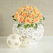This handcrafted composition, prepared with the quiet care of a local Grove Park florist, presents a domed cluster of peach-apricot roses that feel simultaneously sumptuous and restrained. Each rose is carefully selected for its velvety petals and subtle gradient of colour-from creamy peach at the outer edges to warmer apricot tones near the heart-so the bouquet reads as a single, luminous mass. Interspersed among the roses, fine sprays of white gypsophila float like mist, their tiny stars of bloom softening the overall silhouette and catching the light in a way that suggests morning dew. The flowers sit within an open, spherical vessel woven of white wicker; its architectural lattice lifts the blooms, allowing the eye to move freely through negative space while the spherical motif is repeated by smaller decorative orbs beside the arrangement. Fresh green leaves provide cool contrast and depth, their crisp edges grounding the composition and hinting at a subtle, leafy fragrance mingling with the roses' powdery sweetness. Warm daylight-imagine the soft late-morning light that filters through windows around Grove Park station and the Nature Reserve-gently illuminates each petal, creating a tranquil, refined mood. Intended for intimate weddings, milestone anniversaries, or as a gracious house-warming, this arrangement balances sculptural modernity with tender romance. The evident skill in proportion and texture reflects a neighbourhood florist's eye: precise, thoughtful, and attuned to how a bouquet should both complement a home and quietly celebrate life's new beginnings.