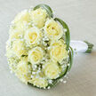 Seen from a slightly elevated viewpoint, this bridal bouquet presents a compact, opulent dome of cream roses-roughly fifteen to twenty blooms-each petal curling into a perfect spiral that catches a soft, early-morning light. Between the larger rose heads, clouds of tiny white Gypsophila lend an airy, dewy texture that contrasts gently with the roses' smooth, satin-like petals. Sprays of bright green grass weave and arch around the bouquet's edge, their slim, glossy ribbons shaping a subtle, natural frame and introducing a contemporary flourish to the otherwise classic composition. At the base, the natural stems are tightly bound in a pristine white satin ribbon finished with a neat knot; the handle looks cool and comfortable to hold, balanced and secure in the hand of a bride leaving for a ceremony near Grove Park's High Street or the quiet lanes toward the local park. The bouquet sits on a light taupe linen background that absorbs light softly and allows faint hints of pale yellow and ivory to glow without glare. The lighting is diffused and flattering, revealing delicate variations in tone and implying the gentle scent of fresh roses mingled with a hint of green. As a locally crafted arrangement by Grove Park Florist, this bouquet reads as trustworthy, refined, and perfectly suited to small neighbourhood weddings, civil ceremonies, or elegant anniversary gifts delivered across Grove Park and surrounding streets.