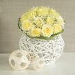 A refined floral centerpiece rests quietly on a pristine white surface, its form defined by a generous dome of light yellow roses that appear to glow softly, as if lit by late afternoon sun filtering through a Grove Park window. Each rose is fully opened, petals layered in rich spirals that touch and overlap, eliminating any harsh gaps and creating a continuous, velvety landscape of colour. Sprinkled through the roses, feathery plumes of white baby's breath add a veil of tiny blossoms, bringing a whisper of texture and a sense of airy lightness to the bouquet. Encasing the base of the flowers is a slim ring of fresh green leaves or delicately flattened foliage, providing a vivid contrast that subtly outlines the shape of the arrangement. The bouquet is nestled into a distinctive white vase, expertly woven from slender strands that echo the look of wicker or rattan, its rounded body narrowing gently at the centre like a modern sculpture. The open weave allows thin beams of light to pass through, which fall as soft, linear shadows across the gleaming tabletop. In front of the vase sit two decorative woven balls, one larger and one smaller, fashioned from pale, natural-looking strips that twist into an open, geometric lattice mirroring the vase's texture. The background is a tranquil blur of light beige, similar to the neutral walls found in many Grove Park homes, ensuring the viewer's attention remains on the interplay of flowers and crafted decor. This arrangement feels perfectly suited to intimate wedding receptions, christening gatherings, or elegant dinner parties along the leafy streets near Grove Park station, bringing with it a sense of calm celebration, thoughtful design, and natural grace.