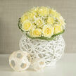 This arrangement reads like a soft composition in ivory and pale yellow: creamy roses arranged into a domed cluster, their ruffled centres unfolding with a gentle confidence, and airy sprays of gypsophila weaving through them like laughter in a quiet room. The bouquet is set upon a white wicker orb that doubles as a tactile sculpture, its open weave adding negative space that allows the blooms to float visually. Under a veil of natural daylight the petals appear almost luminous, and the mingled scent of rose and gypsophila carries a clean, linen-fresh impression tempered by a faint, grounding earthiness-think early summer hedgerows after a gentle rain. Curved stems of greenery loop gracefully around the composition's edges, providing a soft frame and a subtle sense of movement. Minimal woven spheres placed nearby introduce a contemporary playfulness, their matte surfaces echoing the arrangement's modern restraint. The piece feels celebratory yet composed, ideal for marking a thoughtful congratulations, a housewarming along Baring Road, or the centrepiece for a calm gathering in a Grove Park sitting room. As a local florist who often works with neighbours near Chinbrook Meadows and the Grove Park nature reserve, I compose each bouquet to balance scent, texture and line so it reads as both an emotional message and an assured piece of floral design.