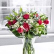 A lush, romantic bouquet of rich red roses, matching red carnations, white alstroemeria and clouds of dainty baby's breath sits proudly in a classic clear glass vase on a bright white table, bathed in soft daylight from a nearby window in Grove Park. The roses are the first thing the eye is drawn to: velvety crimson blooms at different stages, from tightly furled buds suggesting promises yet to unfold, to fully open flowers with generous, layered petals that feel almost touchably soft. Around them, plump carnations in the same deep red tone add fullness and texture, their ruffled edges creating a sense of movement like ripples in fabric. Between the reds, clusters of creamy white alstroemeria, some still closed and others just beginning to trumpet open with faint green-tipped petals, lend a graceful, slightly exotic note. Threaded delicately throughout are tiny white gypsophila blossoms, like small stars or drops of morning mist, softening the contrast between the bolder blooms. The whole arrangement is anchored by abundant greenery: broad, glossy deep green leaves forming a collar at the base, with more slender, lighter stems weaving up through the bouquet to add depth and natural rhythm. The stems are neatly gathered and clearly visible through the transparent vase, immersed in clean water that catches a gentle reflection from the tabletop, subtly reinforcing their freshness as if just prepared for delivery nearby in Grove Park. Behind the bouquet, the window view is intentionally blurred and muted, creating a near monochrome backdrop that makes the reds, whites and greens glow more intensely. The mood is warm, uplifting and elegant, ideal for marking a special birthday, an anniversary, an affectionate thank-you or simply brightening a friend's home on a quiet Chiswick or Grove Park afternoon.