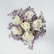 A high-angle overhead photograph shows a compact hand-tied bouquet resting on a pale, almost cloud-grey surface, with no vase in sight so the flowers themselves remain the entire focus. At the centre of the arrangement sit six immaculate white roses, each bloom carefully spiralled, their petals tightly layered yet softly curving at the edges like folded satin. The roses are a creamy ivory white with the faintest hint of green near the sepals, giving them a fresh, just-cut look you might see in a Grove Park florist's cooler early in the morning. Around them billows a generous collar of lilac and muted lavender statice, the tiny clustered flowers forming a misty halo that softens the strong, classic lines of the roses. The purple filler is densely packed but fine in texture, creating the look of delicate lace or heathered foam, and adding depth without overpowering the central blooms. Soft, even lighting from above removes any harsh shadows, allowing each velvety rose petal and each tiny statice floret to be clearly defined, almost touchable. The bouquet is shaped into a near-perfect circle, suggesting a professionally crafted design suited to weddings, quiet anniversaries, or thoughtful sympathy deliveries around Grove Park and the nearby leafy streets. The mood is calm, refined, and slightly introspective, as if the flowers are offering a gentle pause in a busy London day. There are no bold colours or distracting accessories, just the timeless contrast of crisp white and gentle lavender, creating a serene, elegant floral piece that feels both intimate and sophisticated.