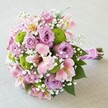 Poised on a gentle, neutral-toned backdrop, this splendid bouquet radiates the refined energy of Grove Park's flourishing gardens. The focal point presents fully opened lavender roses, each exuding softness and romance, their petals closely layered like memories cherished across generations. Scattered amid the central flowers, pink Alstroemeria lilies add splashes of colour-trumpet-shaped petals brushed lightly with yellow and streaked in deep magenta, echoing the vibrant flora of Grove Park's parks. Compact spheres of vivid lime green button chrysanthemums bring excitement and modernity, their spiky texture offering a dynamic contrast that enlivens the bouquet. Between these charming blooms, powdery-white gypsophila threads delicately weave, rendering a gentle, airy touch that softens all edges. Glossy green leaves cradle the composition, while the gathered stems are enclosed in a rich fuchsia ribbon, adorned gracefully with iridescent pearl beads. This bouquet-whether intended for a wedding aisle, a Garden Estate anniversary, or a grand local gesture-captures Grove Park's sense of togetherness and celebration, evoking both elegance and vibrant community spirit.
