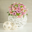 This photograph presents a rounded pastel bouquet that seems tailor-made for a chic Grove Park wedding, carefully styled on a pristine white surface flooded with gentle, natural light. The heart of the arrangement consists of full-bodied roses in a spectrum of lavender and pink, their layered petals opening in generous spirals that create a soft, romantic focal point. Interlaced among them are pale pink alstroemeria, their long petals curving gracefully and adorned with subtle freckles and streaks of deeper colour, giving the bouquet a lively, almost conversational character. Lime green button chrysanthemums pop up throughout the design, their small, domed heads bringing a bright, contemporary edge that contrasts beautifully with the muted pastels and suggests a modern take on classic wedding flowers. Airy sprigs of white baby's breath thread delicately through the composition, providing a misty, ethereal fill that softens lines and adds volume without heaviness. Beneath the blossoms, glimpses of rich green foliage provide structure and a quiet, natural frame. The entire floral dome is presented in a white, globe-shaped vase crafted from intricately woven wicker or rattan, the open lattice pattern forming an eye-catching sculptural base. Two smaller, natural-toned woven spheres sit close by on the same white surface, picking up the texture of the vase while offering a more rustic, relaxed feel reminiscent of stylish homes near the Thames in Grove Park. A calm, neutral wall in the background ensures there are no distractions from the artistry of the flowers. The overall mood is hopeful and refined, perfect for bridal collections, anniversary surprises, or elegant dinner parties in and around Grove Park.