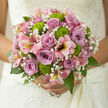 The image shows a romantic hand-tied bouquet, its palette of lilac, pink, and fresh green arranged with the restraint and beauty of a Grove Park garden at first light. At the heart of the bouquet sit lilac and lavender roses, their petals velvety and cool, spiralling inward with precise grace, each bloom suggesting patience, affection, and trust. Between these roses, fine sprays of gypsophila form a delicate, cloud-like backdrop, their tiny white blossoms adding depth and a gentle softness that wraps around the focal flowers. Bright green button chrysanthemums pop from within the design, their rounded shapes and densely layered petals bringing a lively contrast, as if echoing the vibrant lawns and hedges visible along the nearby Strand-on-the-Green. Pink alstroemeria extend outward in gentle arcs, their long, slightly curved petals brushed with strokes of warm yellow and rose, creating a sense of movement that guides the eye around the bouquet. Encircling the flowers, glossy green foliage provides a lush frame, leaves overlapping in varying shades to create texture and natural volume without feeling heavy. The light that falls across this arrangement is subtle and even, casting soft shadows and enhancing the pastel tones, giving the impression of a bouquet placed near a window in a quiet Grove Park home just before being carried out to a local ceremony. Everything about the composition suggests thoughtful craftsmanship, from the balance of colours to the interplay of textures, resulting in a bouquet that feels both refined and approachable, ideal for vows whispered in familiar, much-loved surroundings.