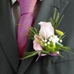 A moment of joy and artistry is distilled in this close-up image, capturing the refined ambiance of a Grove Park celebration. The sharply pressed charcoal suit jacket sets a distinguished stage, while a magenta-pink silk tie brings a vibrant flash of color beneath. The visual focus, however, is drawn instantly to the lovely boutonniere pinned to the left lapel-a soft pink rose at its heart, its petals blushing and inviting, surrounded by a pink alstroemeria flecked with violet, echoing the playful side of Grove Park's community spirit. Encircling these blooms are clusters of fluffy white gypsophila, casting a gentle haze like early-morning mist over Dukes Meadows. The brilliant green button chrysanthemum pops with life and energy, embraced by feathery fern sprigs and graceful green blades that add depth to the display. An iridescent lavender ribbon bow sits neatly at the base, mirroring the tie for a harmonious finish. Designed by skilled local florists and photographed in the warm, natural light synonymous with Grove Park, this boutonniere embodies the hopes, excitement, and elegant traditions that define weddings and essential gatherings throughout the neighbourhood.