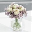 A close-up photograph captures a tranquil bouquet of creamy white roses nestled into a soft cloud of lavender-hued statice, arranged with quiet precision by a local Grove Park florist. Each rose is fully formed yet still fresh, with velvety petals gently unfurling from a pale, buttery centre into ivory outer edges, creating a subtle gradient that conveys purity and calm. The roses sit slightly elevated above the surrounding statice, so their rounded, classic blooms appear to float over a delicate haze of tiny, papery purple flowers. The statice is airy but abundant, its many branching stems weaving in and out around the central roses, adding texture and a slightly rustic, countryside feel that would be at home along the leafy streets near Grove Park station. All the stems are gathered into a clear glass vase with a distinctive crackled, ice-effect surface; the textured glass bends and scatters the light, forming tiny glimmers that echo the bouquet's gentle sparkle. Through the water inside the vase, the green rose and statice stems remain clearly visible, crossing and layering in natural lines that emphasise the hand-crafted character of the design. The arrangement stands on a smooth, bright white surface that reflects just enough light to create soft, feathered shadows under the vase, while the background fades into a pale grey blur that keeps every ounce of attention on the blooms. The overall atmosphere is one of delicate tranquillity, like a quiet afternoon along the paths by the River Thames near Grove Park, making this arrangement ideal for a soothing birthday gesture, a thoughtful sympathy gift, or simply to bring a sense of serene elegance to a peaceful corner at home or in a calm workspace.
