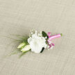 This image shows a thoughtfully composed boutonniere resting on a pale, textured background that recalls soft woven fabric, giving the whole scene a calm, tactile warmth. The focal point is a fully open white lisianthus, its petals layered and gently ruffled, forming a generous bloom that seems to float above the surface. The creamy white has a subtle richness that suggests luxury without ever feeling showy. Snuggled up against this central flower are two lisianthus buds, still mostly closed, their smooth, ivory-cream forms offering a quiet counterpoint to the exuberance of the open bloom, like a promise yet to be fulfilled. Around and between them, small clusters of baby's breath scatter in delicate sprigs, each cluster made up of tiny, snow-white flowers that add an ethereal, lace-like quality to the design. Fine green foliage and slender stems stretch outward, adding freshness and a sense of movement, as if a breeze from nearby Grove Park station had stirred them just moments before. The lower portion of the boutonniere is tightly wrapped in a bright pink satin ribbon, whose vivid colour and glossy texture introduce a playful, contemporary note. The ribbon winds precisely around the stems and is finished with a petite bow, its loops catching soft highlights and lending a polished, celebratory air. Overall, the colour palette of white, green, and bold pink makes this boutonniere particularly fitting for weddings, proms, or intimate celebrations in and around Grove Park, providing a small but expressive floral accent that speaks of care, joy, and the crafting hands of an experienced local florist.