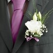 A close-up photograph captures the left lapel of a groom's dark charcoal grey suit, the fine wool cloth showing a subtle woven texture that sits smoothly against a crisp, patterned white and grey shirt. At the centre of the frame, a richly coloured magenta silk tie, expertly knotted, glows with a soft sheen, its saturated hue immediately drawing the eye and setting a confident, celebratory tone. Pinned neatly to the lapel is a carefully crafted buttonhole featuring a single, fully open white lisianthus bloom, its many ruffled petals layered like soft satin, each edge gently curling to create a cloud of creamy white. Just above the main flower, two slim ivory buds rest closely together, suggesting blossoms still to come, while tiny stars of baby's breath spill lightly around them, forming a delicate halo of miniature white flowers. Wisps of fine greenery arc out in slender lines, adding freshness and movement, and a broad green leaf curves protectively at the base, cradling the stems. A narrow mauve ribbon, tied with precision, wraps the buttonhole, echoing the pink-toned tie and uniting the colours into one harmonious design. The lighting is gentle and even, as if the groom is standing near a window above Grove Park station, ready to step out for a local ceremony. The overall mood is one of quiet anticipation and polished elegance, the sort of refined detail a Grove Park groom might choose for a wedding in a nearby church or a sophisticated celebration close to the tree-lined streets and calm suburban charm of south-east London.