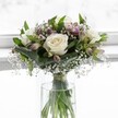 An elegant white rose bouquet stands in a simple cylindrical glass vase, bathed in soft daylight that feels like it could be streaming through a bright Grove Park bay window. Three creamy white roses sit at the heart of the design, fully open yet still perfectly fresh, their velvety petals curving outwards to reveal warm ivory centres that suggest purity and calm. Around them, deep green foliage forms a gentle cradle: some leaves are broad and slightly folded, others slimmer and more upright, all adding layers of rich emerald texture and subtle shadows that shape the rounded, compact form of the arrangement. Threaded delicately between the roses and greenery are wisps of tiny white gypsophila, or baby's breath, like light clouds or morning mist, giving the bouquet a soft, romantic haze. Slim alstroemeria buds and blooms in muted mauve and blush-pink peek through the foliage, their petals only partially unfurled, adding quiet hints of colour and the promise of further blossoming. The stems, visible through the clear water of the vase, are neatly gathered and secured with a fine ribbon just above the waterline, showing the careful hand of an experienced florist. The background is a luminous, softly blurred white, with no distractions, allowing every petal, leaf, and stem to be clearly defined in the glow. The mood is tranquil and reflective, ideal for a gentle condolence gesture, a serene wedding morning in a Grove Park townhouse, or a thoughtful token of support delivered to a friend across the neighbourhood, radiating understated grace and natural harmony.