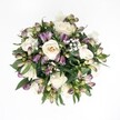 Top-down view of a lush, circular bouquet resting against a crisp white background, its colours standing out as clearly as a map of Grove Park on a bright morning. At the heart of the arrangement, creamy white roses in shades of ivory and soft champagne open in layered spirals, some fully unfurled and plush, others still cupped and coy, as if just beginning to tell their story. Threaded between them are deep lavender and rich purple alstroemeria buds, their slender petals streaked with darker tones and fading to fresh green at the base, adding rhythm and movement to the design. Fine clouds of white gypsophila drift through the bouquet like a soft mist over the trees along Marvels Lane, lending an airy, almost weightless quality. Dark green foliage, broad and glossy, cradles the blooms from beneath and peeks through between petals, creating a rich, natural frame that makes the lighter flowers glow. The lighting is gentle and even, with no harsh shadows, as though the bouquet were laid under a skylight in a quiet Grove Park kitchen. Every element appears freshly gathered, the roses velvety and cool, the alstroemeria buds firm and promising, the greenery crisp to the touch. The mood is calm and deeply romantic, suitable for a local anniversary surprise, a wedding morning in SE12, or a thoughtful gesture of comfort. This arrangement feels like a quiet promise: elegant, composed, and full of unspoken affection, a timeless floral embrace captured from above in serene, modern simplicity.