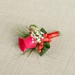 A richly detailed boutonniere is laid carefully on a softly textured, light beige linen background, the kind of woven fabric you might see on a table in a Grove Park townhouse. At the heart of this small floral piece is a single deep fuchsia rose, its velvety petals tightly coiled at the centre and gently unfurling towards the edges, suggesting a bloom cut at just the right moment of freshness. Around the base of the rose, a halo of airy white baby's breath creates a delicate, cloud-like contrast, each tiny blossom catching the soft, natural light that falls across the scene. Threaded between these white clusters are plump, glossy red hypericum berries, their rounded forms providing a joyful spark of colour and a hint of festive charm, as if ready for a winter wedding at a local Grove Park church. Wisps of fine, fern-like greenery stretch out from the composition, adding movement and a slightly wild, garden-picked feel, while broader, richer green leaves are folded and curled with intention, giving the design structure and a sculpted elegance. The stems are neatly bound together with a rich red satin ribbon, wrapped smoothly and finished in a neat bow that echoes the romantic tones of the rose. Viewed from a slightly raised angle, every detail of the craftsmanship is visible: the way the foliage frames the bloom, the way the berries nestle close, and the balance of colour and texture. The boutonniere feels fitting for a groom or groomsman on a Grove Park celebration, capturing the emotion of the day in one small, artful floral accent, ready to be pinned to a lapel before stepping out into the streets around the local park or station.