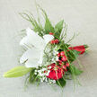 Seen up close, this bouquet looks like a small, hand-held story crafted by an experienced Grove Park florist, perfect for a wedding, engagement, or romantic surprise. The central white lily is fully open, its broad petals arching outwards with poised elegance, their surface satin-smooth and almost luminous against the surrounding colour. Within its centre, a soft brush of golden pollen adds a touch of warmth, drawing the eye inward. Around the lily, rich crimson roses cluster together, their velvety petals unfolding in dense layers that suggest devotion and heartfelt emotion-like a quiet vow exchanged in a tucked-away corner of Grove Park. Between these roses, glossy red berries peek out, their bright tones and rounded shapes contributing energy and a hint of festive charm. Wisps of white gypsophila weave delicately among the larger blooms, the tiny flowers creating a fine, cloud-like veil that softens the bold reds and adds an air of innocence. Light, feathery foliage curves around and between the flowers, bringing a sense of movement and natural rhythm, as if a gentle breeze has passed through the bouquet. The stems are neatly gathered and tightly wrapped in a narrow strip of red satin ribbon, smooth to the touch and visually striking, giving the design a refined, ceremonial feel. The overall impression is one of elegant warmth and quietly confident romance, an arrangement that would look just as fitting in a bridal portrait on Burlington Lane as it would arriving by same-day delivery to a doorstep in Grove Park, carrying with it the message, You matter deeply to me.