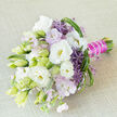 On a pale, textured beige fabric that looks like finely woven linen, a hand-tied floral bouquet is placed on a gentle diagonal, as if just set down by a bride pausing for a moment in Grove Park. The composition is a thoughtful harmony of white, lavender, purple, and fresh green, the colours working together in a calm yet sophisticated palette. Full, cloudlike white lisianthus flowers dominate, their soft, layered petals curling back to reveal subtle creamy yellow centres, while neighbouring stems hold lisianthus buds in pale green and off-white, poised to open. Nestled between these are clusters of delicate lavender-pink blossoms that introduce a dreamy, pastel mist throughout the bouquet. Punctuating this softness are vivid purple allium florets, their star-shaped forms bursting outward like tiny fireworks, adding structure and a touch of playful modernity. Surrounding and threading through the flowers is a generous selection of greenery: elongated leaves, lighter green spiky elements, and loops of grass-like foliage that provide texture, depth, and movement. The stems are tightly gathered and wrapped in a bold fuchsia satin ribbon, its sheen catching the light, then finished with an elegantly draped strand of small white pearls that encircle the handle like a bracelet. The overall effect feels perfect for a wedding at a local church near Grove Park or a refined celebration in a nearby garden, conveying purity, romance, and joy. As experienced florists might describe it, this bouquet balances classic bridal styling with a chic, contemporary twist, suggesting soft fragrance, heartfelt emotion, and the quiet luxury of expertly arranged fresh flowers delivered straight from a trusted local studio.