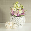 An artfully styled floral arrangement rests on a smooth white surface, its colours and textures combining in a way that feels both modern and warmly familiar, like a carefully curated corner of a Grove Park living room. At the centre is a lush, dome-shaped bouquet of lisianthus in creamy white and soft buttery tones, their many layered petals opening in gentle spirals. Around them, clusters of delicate pink stock create a cloud-like softness, while scattered purple allium blooms introduce small bursts of spiky, architectural interest. A collar of rich green leaves wraps around the base, giving the flowers a tailored finish that hints at the steady hand of an experienced florist. The whole bouquet is gathered into a distinctive spherical vase formed from interwoven white twigs, creating a sculptural, open lattice that allows light to pass through and reveals glimpses of the stems inside. Beside the vase on the white tabletop lie two decorative wicker spheres in pale, natural wood, their crisscrossing strips echoing the woven structure of the vase and tying the scene together. The background is softly out of focus, a neutral greige wall with a subtle texture that suggests fabric or finely brushed plaster, offering a calm, sophisticated backdrop. The lighting is even and gentle, with no harsh shadows, so the pastels appear tender and fresh. This is the kind of centerpiece that would sit comfortably on a Grove Park dining table for a relaxed Sunday lunch, or on a console in a local family home, quietly celebrating spring, new beginnings, or a thoughtful thank-you without overwhelming the space.
