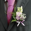 A tightly framed close-up shows the upper torso of a man dressed for a wedding in Grove Park, his charcoal suit jacket and pale patterned shirt forming a refined backdrop for an exquisite buttonhole. The soft grid of the light shirt peeks out behind a bold magenta silk tie, its knot perfectly centred and its surface catching the gentle daylight like a ribbon of colour running down the chest. Pinned with care to the left lapel, the boutonnière becomes the true focal point: a small, sculpted garden of pastel blooms and greenery that feels freshly gathered from a quiet corner of nearby Grove Park itself. At the top, slender, unopened buds of creamy white flowers, similar to snapdragons, lift the eye upwards, their smooth, tapered forms suggesting blooms still on the cusp of opening. Nestled beneath are clusters of soft pink and lavender-purple petals, reminiscent of sweet peas or delicate stock, their ruffled edges layered like tissue-thin silk. Tiny stars of white gypsophila drift between them, creating a misty, cloud-like texture that lightens the whole composition, while a few deeper purple florets add just enough depth and contrast to keep the palette interesting without overpowering the subtle romance. Broad, glossy dark green leaves cradle the flowers, offering structure and polish, while fine, feathery fronds of bright green foliage arc gently outwards, lending natural movement and an almost woodland softness. Around the stems, a satin ribbon in muted lavender is tied into a neat, symmetrical bow, echoing the tie's vibrancy but with a quieter, more romantic tone. The overall impression is of a groom or honoured guest caught in a candid, serene moment before the ceremony, his flowers and attire harmonised with understated elegance for a Grove Park celebration.