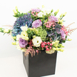 A contemporary celebration of abundant colour and form, this luxury bouquet from Grove Park Florist is arranged in a matte black square box, its clean lines a striking contrast to the organic exuberance within. At the heart of the design are voluminous clusters of deep blue and periwinkle hydrangeas, their pillowy textures forming a tranquil base reminiscent of the Thames at dusk. Interwoven are roses in soft lavender and blushing pink, their petals elegantly opening to convey affection and grace. Creamy white lisianthus, some tinged gently with pale yellow, provide brightness and classical charm, while vivid lime-green filler, likely 'Green Trick' dianthus, and delicate sprigs of pink astilbe introduce playful texture and a whisper of wild meadow. Glossy greenery and emerging flower buds cluster at the edges, lending a lush, fresh-from-the-garden appearance evocative of Grove Park's flourishing borders and vibrant street markets. Every colour is heightened under clear natural light, and the bouquet's rounded form is expertly composed to be admired from every perspective. This arrangement is perfectly suited for celebrations or acts of appreciation throughout Grove Park, designed to bring local floral beauty and an air of sophisticated joy into any beloved space.