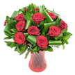 A luxurious, rounded bouquet of a dozen or more deeply saturated crimson roses fills the frame, each bloom opened to reveal tightly spiralled, velvety petals that catch a soft, even studio light. The petals read almost wine-dark at their centres and open to a luminous scarlet at the edges, their texture giving a tactile sense of plushness and warmth. Between the rose heads a generous sweep of glossy, dark green foliage provides volume and contrast: dense, flat leaves-reminiscent of Ruscus or Salal-form a verdant backdrop while broader, emerald Aspidistra-like leaves are carefully rolled and curled into sculptural loops and spirals. Those dramatic green curves create an architectural rhythm around the roses, cradling blooms and guiding the eye in vertical and circular lines. The stems sit in a tall, slender ruby-red glass vase with a subtle ribbed or twisted surface that catches light in vertical streaks; the translucency of the glass deepens the bouquet's palette and reinforces the arrangement's romantic mood. Everything is set against a pristine white background so the red and green leap forward with clarity, isolated as a single striking focal point. The composition feels timeless and intimate - a classic romantic statement that also reads as contemporary design because of the folded foliage and sculpted form. I imagine the faint, classic scent of fresh roses rising from the cluster, an aroma that would suit an anniversary table, a heartfelt Valentine's delivery, or an elegant centerpiece for a candlelit dinner. Locally, this is the kind of arrangement you might choose for a celebration near Grove Park green spaces, Hither Green lanes, or a quiet supper close to Lee's neighbourhood cafés.