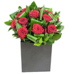 A compact, modern composition that pairs sumptuous crimson roses with crisply folded emerald foliage, presented as if photographed on a bright morning in Grove Park. Each rose opens in velvety spirals, the deep red petals catching the light with a soft, velvet sheen while the surrounding leaves curl and overlap like green ribbons, their glossy surfaces reflecting highlights from a clear, contemporary studio lamp. The blooms sit low and close together, their rounded heads forming a dense, emotive cluster that reads as both intimate and assertive. A matte black square cube anchors the design, its geometric silhouette giving weight and contrast to the lush, organic forms above and lending the whole piece an urban-chic edge suited to small townhouses or city flats around the station. Running a fingertip near the arrangement, you would sense the difference between the rose petals' silk-like softness and the leaves' firmer, almost waxy texture; inhale and the heady, classic scent of fresh roses drifts through a cool green top-note from the foliage. The florist's hand is visible in the rhythmic placement of each leaf, looped and tucked to create movement without clutter, a technique I often use for deliveries to local addresses around Chinbrook Meadows and nearby roads. This bouquet feels designed for quiet declarations-a meaningful anniversary gift, a romantic milestone, or a thoughtful gesture to brighten a bedside table. It reads as cultivated emotion in a contemporary frame, a present that looks as considered as it feels, made to be noticed, embraced, and remembered.