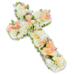 A softly coloured funeral flower cross is set against a pure white background, its gentle hues and intricate detailing offering a sense of stillness and consolation. The cross itself is completely cloaked in flowers, giving it a thick, substantial profile and a velvety, cushioned texture. Dominating the arrangement are white and ivory blossoms, including what appear to be hydrangea heads, spray roses, and other compact flowers, all tightly woven together to form the base of the Christian symbol. Nestled among this white foundation are scattered clusters of pastel roses in shades of pale peach, apricot, and blush pink, which add warmth and dimension. These roses, some fully opened and others delicately budding, create soft focal points as the eye travels from the centre to the arms and down the length of the cross. Small flecks of lavender or light purple filler flowers peek out here and there, like whispered notes of colour that enhance rather than interrupt the serene palette. Slim strands of green foliage appear around the edges and occasionally between blooms, outlining the cross with a fresh, natural frame. The lighting is bright and even, producing a faint shadow underneath that gives the impression the floral cross is gently raised from the surface, much as it might be elevated on a stand in a Grove Park church or chapel. This type of floral tribute, often chosen for funerals, memorials, or graveside services in the local area, quietly symbolises faith, remembrance, and the hope of peace, providing grieving families with a beautiful, respectful way to say a final farewell.