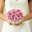 There's a quiet, almost reverent beauty in this moment, where a bride's hands encircle a bouquet alive with energy and hope. In the dappled light familiar to Grove Park's tree-lined lanes, plump pink roses stand at their peak, their petals swirling from gentle blush hearts to bold, fuchsia-tinged edges-the very image of romance and promise. Threads of baby's breath are woven between the blooms, adding frothy brightness and an airiness that floats softly above her dress's intricate lacework. The delicate polish on her ring finger glints in the light, and the gown's gathered sash drapes gracefully above her hands, hinting at movement and subtle joy. The whole scene, softened by a muted background, embodies the anticipation and happiness of a new beginning. It is a reflection of Grove Park Florist's commitment to crafting wedding florals that capture both the grandeur and the gentle intimacy of a romantic local ceremony.