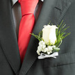 In the soft studio light, an ivory rose boutonnière makes a considered statement against matte charcoal tailoring, the bloom's velvety layers opening in a way that feels both tender and assured. Clustered at the base, delicate baby's breath provides a frothy halo of tiny white puncta that temper the rose's form with innocence and breathy texture, while fine slivers of fern and twisted leaves reach up in graceful arcs to add movement and dark-green depth. The boutonnière is pinned just above the line of a smart checked shirt and beside a bold red silk tie, the contrast of textures - wool, cotton, silk, and soft petal - creating a tactile dialogue that reads as timeless romance. A neat white ribbon bow secures the stems, its satin sheen catching a hint of reflected light and lending the piece a ceremonious finish. The overall palette is cool and composed: soft creams, deep greens, and charcoal, with the red tie offering a vivid punctuation. You can imagine the faint, comforting scent of rose and green leaves, like an early-summer morning near Grove Park station or a walk through the local nature reserve before a ceremony. Crafted with steady hands and an eye for proportion, this boutonnière is intended for a groom or someone special - an intimate accent that complements formalwear without stealing the scene, quietly marking the day's importance with graceful simplicity.