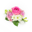 Set against a pristine white background, this compact corsage reads like a small, personal love letter. At its centre sits a plush, bubblegum-fuchsia rose whose young petals curve inward in a velvet spiral, then unfurl toward the edges with richness and depth. Surrounding the rose, clusters of pale pink to lavender hydrangea petals create a soft, billowing cushion - airy and almost tactile - that softens the rose's intensity with romantic pastels. Scattered among these are creamy white, trumpet-formed blooms with a faint green throat, their smooth, satiny surfaces catching the light and offering a gentle, pure contrast. Glossy, broad green leaves tuck in at the base, their deep emerald sheen grounding the piece and carrying a fresh, leafy scent when handled. Slim, upward-reaching green buds tipped with a whisper of yellow add a hopeful, living momentum, as if this little arrangement promises more blossoms to come. A tiny cluster of pearl-like beads cascades at the lower right, luminous and warm against the florals, suggesting bridal heirlooms or a prom corsage with a classic twist. The whole styling is lit in soft, natural daylight that emphasizes texture - the rose's velvet, the hydrangea's papery layers, the pearls' satiny gleam - evoking a gentle, celebratory mood suited to weddings, anniversaries, or intimate ceremonies. As an arrangement a florist would tuck into a sash or pin to a lapel, it speaks of careful handcraft, seasonal freshness, and quiet luxury - perfect for spring and summer events around Grove Park and neighbouring South East London communities looking for something both refined and tender.