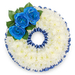 A bespoke sympathy tribute formed in the distinct shape of the number '0', photographed from above against a clean white background. The numeral is densely covered with hundreds of small white spray chrysanthemums, their tightly packed florets creating a pillowy, tactile surface that reads as quiet and pure-soft to the eye, subtly luminous in the light. Around both the outer and inner rims of the '0' a pleated royal blue satin trim follows the contour, its ruffled scallops echoing the curve of the number and lending a formal, refined border that matches the colour of the accent blooms. On the upper left curve sits a careful cluster of three royal blue roses, their deep, velvety petals unfurling from compact centres; a few dark green glossy leaves nestle behind them, offering a cool, natural contrast to the blue and white palette. The blue here feels intentional and personal-serene as a twilight sky, dignified as a uniform or favourite shade chosen to honour someone specific-and the ribbon's sheen catches the light with a gentle, respectful gleam. The chrysanthemums' faint, clean scent and the satin's subtle rustle combine into a composed sensory whole, one meant to console rather than overwhelm. Designed as part of a custom numbered tribute for memorial services, this '0' can represent an age, an anniversary, or a meaningful numeral in a loved one's life; its balanced geometry and restrained colours make it suitable for a chapel or funeral parlour. As a local florist we craft these with steady hands and attention to symbolism, mindful of the way colour and form can carry memory and comfort across the first hush of a farewell.