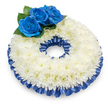 Photographed from a slightly elevated angle against a clean white backdrop, this circular sympathy wreath reads as a quiet, comforting presence, its soft shadow anchoring the form on the surface. The body of the wreath is a dense, almost cloud-like band of pristine white chrysanthemums; their feathery petals overlap in hundreds of tiny layers, the centers carrying a faint creamy-yellow warmth that prevents the white from feeling stark. At the upper-left quadrant, three royal-blue roses form a gentle focal cluster - their velvety, tightly spiralled petals glow with a deep sapphire intensity and are cushioned by rounded, dark green foliage that brings a natural, living contrast. Tracing both the outer and inner edges of the chrysanthemum ring is a pleated satin ribbon in the same deep cobalt hue, its glossy ruffles catching soft highlights and adding a formal frame to the piece. The lighting is soft and even, revealing texture and sheen rather than strong shadows, and there is a subtle, comforting scent suggested by the image: the clean sweetness of chrysanthemums balanced by the faint, green aroma of foliage. As a florist familiar with Grove Park and nearby community spaces like Grove Park Nature Reserve and the streets around the station, I find this palette especially suited to memorial services and intimate remembrances - dignified, unobtrusive, and personal. The composition speaks of peace and steady support, a respectful tribute that would sit calmly beside a pulpit or on a memorial table. For local requests, similar wreaths can be prepared with care for services across Grove Park and neighbouring Lewisham areas.