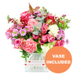 The image captures a generous, garden-style bouquet arranged in a contemporary square glass vase, set against a clean, white studio backdrop that highlights every nuance of colour. The design centres around an array of pink roses, from rich fuchsia and bold magenta to tender blush shades, each bloom at a slightly different stage of opening, which gives the piece a sense of life and unfolding emotion. Nestled amongst them are lush carnations in coral, peach-pink, and soft pastels, their densely ruffled petals providing a plush, textured counterpoint to the smooth rose petals. Interwoven through the arrangement are clusters of daisy-like chrysanthemums, tinted in lavender and light purple tones, their bright green centres adding a refreshing burst that draws the gaze across the bouquet. Spots of creamy white flowers are scattered with care, adding lightness and allowing the colours to breathe. Surrounding the flowers is a collar of greenery: rounded eucalyptus leaves with a delicate, silvery-blue cast, darker glossy foliage, and a few reddish-brown accents that subtly suggest warmth and intimacy. The stems meet in a tidy bundle inside the clear glass vase, whose base is wrapped with a white band decorated with repeating red hearts and the words all my love written in a flowing script, reinforcing the romantic, heartfelt nature of the gift. In the bottom right corner, an orange circular label with crisp white capital lettering declares VASE INCLUDED, signalling that the display arrives exactly as pictured. The bright, even lighting suggests a bouquet freshly delivered from a Grove Park florist, ready to be placed on a hallway table, beside a window looking toward the park, or on a breakfast tray for a special occasion such as Mother's Day, a birthday, or a just-because surprise.
