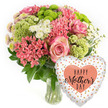 The scene shows a bright, affectionate Mother's Day gift display: a lush floral bouquet in a glass vase accompanied by a heart-shaped foil balloon, all set before a seamless white background. The bouquet is confidently rounded, filling the frame with colour. Several large pink roses crown the arrangement, each rose open enough to reveal its layered petals, edged with a deeper, more saturated pink that adds depth and romance. Nestled alongside are tightly clustered coral-pink flowers, similar to kalanchoe, creating textured islands of small, many-petalled blooms. Lime-green button chrysanthemums dot the design, their neat, spherical shapes and delicate, finely fringed petals lending a contemporary, playful edge that contrasts with the traditional charm of the roses. Interwoven throughout the bouquet are dainty white spray chrysanthemums with daisy-like faces, their bright centres and crisp petals introducing pockets of light that echo the clean background. The entire mix is supported by rich, dark green foliage, leaves curving naturally to provide a structured base and a sense of fullness that suggests a skilled florist's hand. The stems, visible through the transparent vase, offer a natural vertical rhythm and hint at the freshness of the flowers, ready for delivery to homes around Grove Park and the neighbouring streets of Chiswick. To the right of the vase, slightly lower, rests a gleaming heart-shaped foil balloon. Its pale surface is patterned with random metallic gold and peach dots, giving a festive, confetti-like feel. In the middle sits a bold peach heart with the words HAPPY Mother's DAY picked out in elegant black lettering. Together, bouquet and balloon create a visual message of love and appreciation, ideal for making Mother's Day feel special in Grove Park.