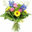 A colourful, garden-style bouquet is displayed upright against a bright white background, like a freshly made arrangement waiting on the counter of a local Grove Park florist. At the focal point, several fully opened pink roses bloom in soft, layered spirals, their velvety petals capturing light and offering a sense of tenderness and warmth. Around them, bold yellow gerbera daisies with rich dark brown centres create a lively ring of colour, their daisy-like faces adding a playful note and a strong visual contrast. Rising above the central flowers, tall stems of rich blue delphinium lift the eye upwards; each stem carries a column of small florets, bringing an elegant vertical line and cool blue tones reminiscent of the Thames in the late afternoon. The bouquet is generously framed with greenery: fine, feathery fern fronds trail from the lower edges and sides, adding softness and movement, while larger, glossy deep green leaves, some showing subtle lighter stripes, have been carefully wrapped around the base of the blooms to form a smooth, architectural collar. Between the more prominent flowers, bright green, cushion-like filler foliage adds texture and volume, giving the bouquet a full, abundant appearance like a well-tended garden near Chiswick or Strand on the Green. A few slender, pale green stems tipped with tiny buds extend lightly beyond the main outline, contributing an airy, freshly picked feel. Below, the stems are neatly cut, aligned, and bound with a simple, natural-looking tie, emphasising the hand-tied, artisan character of the arrangement. The overall mood is uplifting and versatile, perfect for birthdays, congratulations, or offering a thoughtful gesture to someone in Grove Park.